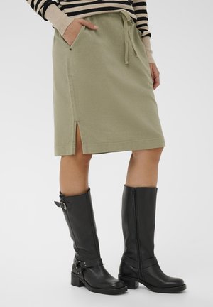 Kobieta ubrana w oliwkową spódnicę do kolan z bocznym rozcięciem, czarne skórzane buty do połowy łydki oraz pasiasty top z długim rękawem i beżowymi mankietami.