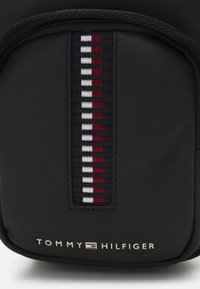 Bolsa de nylon negra con una franja vertical que presenta secciones en rojo, blanco y azul marino, y un logotipo en relieve de "TOMMY HILFIGER" en la parte inferior.