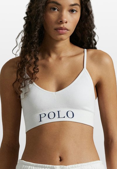 Bílá bavlněná braletka s výstřihem do V a tenkými ramínky. Na pásku je námořnicky modré logo "POLO". Hladká textura, minimalistický styl.