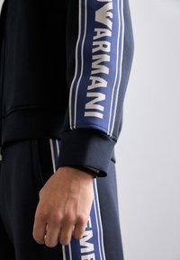 Suurendatud vaade käele ja käele, mis kannab tumesinist riietust, millel on valge tekst "NAVARANI" sinistes triibulistes kangast paneelides.