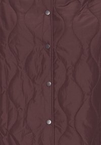 Vêtement d'extérieur matelassé de couleur bordeaux riche avec des motifs de couture ondulés et des boutons-pression en métal noir le long du centre avant.