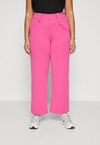 Person klädd i ljusrosa högmidjade vida byxor i kombination med vita och gula Nike-sneakers, som står framför en neutral bakgrund.