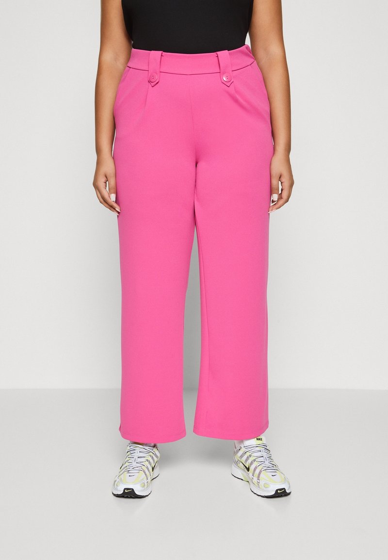 Person klädd i ljusrosa högmidjade vida byxor i kombination med vita och gula Nike-sneakers, som står framför en neutral bakgrund.