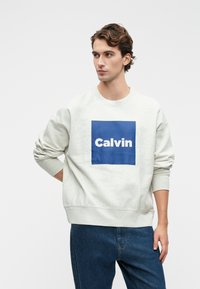 Mladý muž s kučeravými hnedými vlasmi, oblečený v bielej mikine s modrým štvorcom a textom "Calvin", v kombinácii s modrými džínami, stojí na bielom pozadí.