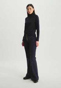 Haut à col roulé bleu marine associé à un pantalon en velours côtelé évasé avec cinq grands boutons sur le devant. Texture lisse et design ajusté.