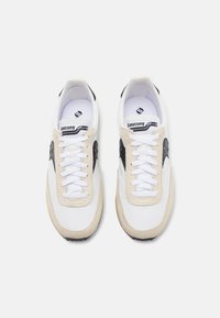 Hvide og beige sneakers med sorte accenter, overdækning i stof, snørebåndslukning og struktureret gummisål. Saucony-logo på tungen og siderne.