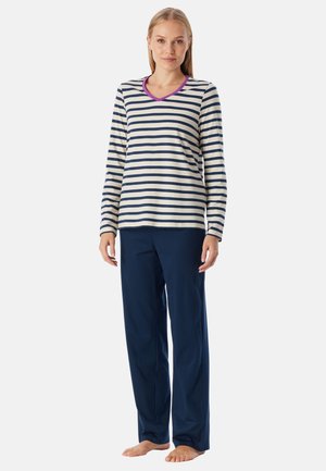 Vrouw die blootsvoets staat, gekleed in een langemouwen crème- en marineblauw gestreept shirt met paarse rand en losse marineblauwe broek.