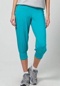 Pantalons courts turquoise avec une texture lisse, dotés de poignets élastiques et d'une coupe décontractée. Associés à des chaussures de sport grises.