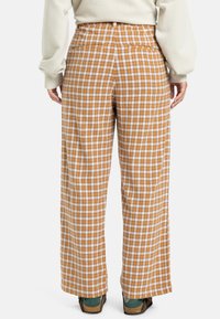 Brede, hoog-waist broeken in een bruine ruit met witte accenten. Bevat twee achterzakken en heeft een gladde stofstructuur.