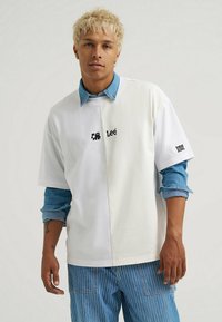 Biała, oversize koszula z denimowym kołnierzykiem, z logo "Lee" i akcentami. Noszona na niebieskich, paskowanych spodniach, ukazujących kontrastujące tekstury.