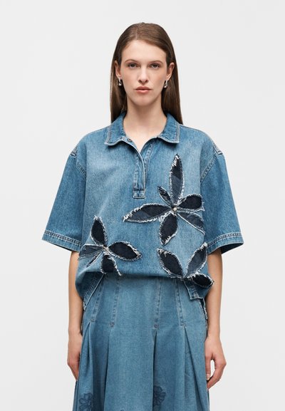 Πολό κοντομάνικο από τζιν με κεντημένα λουλουδάτα διακοσμητικά από σκουρόχρωμο τζιν, ξεφτισμένες άκρες και άνετη γραμμή. Συνδυάζεται με φούστα από το ίδιο ύφασμα τζιν.