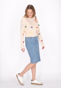 Cardigan crem cu aplicații în formă de fundițe colorate, asortat cu o fustă din denim și adidași albi cu accente gri și negre.