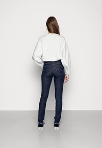 Sudadera gris claro de corte holgado, jeans skinny azul oscuro y zapatos negros sin cordones con acentos coloridos, vistos desde atrás.