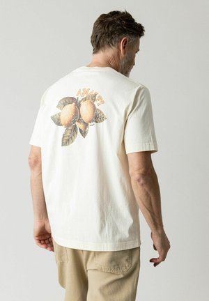 Uomo che indossa una maglietta color crema con un'illustrazione di tre pesche e foglie e il testo "AMAZE" sulla schiena, abbinata a pantaloni beige.