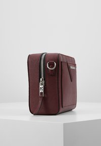 Burgundowa torebka typu crossbody z teksturowanego materiału syntetycznego, z metalowymi zamkami w kolorze srebrnym i bocznym uchwytem na ring. Prostokątny kształt z kieszenią.