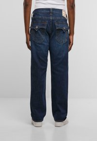 Temno modri kavbojki z ohlapnim krojem, ki imata dve zadnji žepki z značilnim šivanjem in kovinskimi detajli ter oznako na pasu.