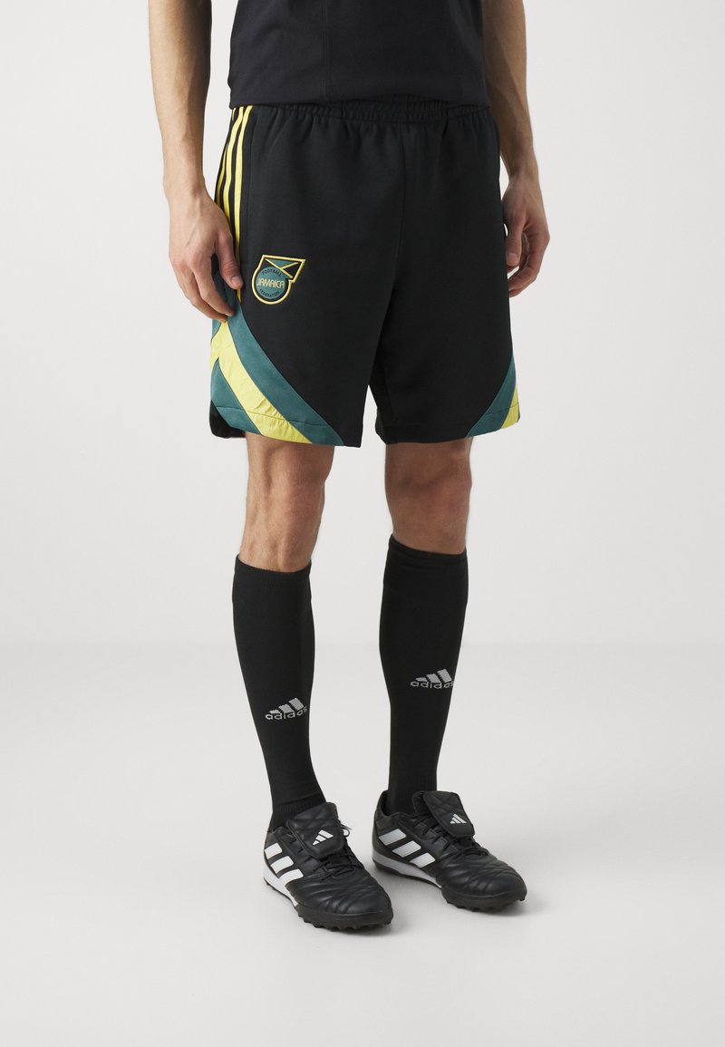 Pantalones cortos de fútbol negros con acentos en amarillo y azul verdoso, que presentan un logo circular de "Jamaica". Combinados con calcetas negras y zapatillas deportivas negras con franjas blancas.
