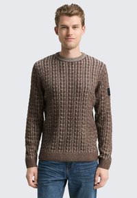 Brauner Strickpullover mit strukturierten vertikalen Streifen, Rundhalsausschnitt, gerippten Bündchen und einem Markenpatch am linken Ärmel. Getragen mit blauen Jeans.