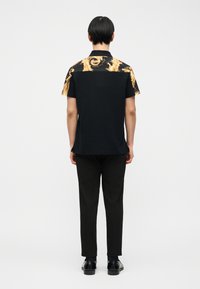 Άνδρας που φοράει μαύρο παντελόνι και μαύρο κοντομάνικο πουκάμισο με χρυσή διακοσμητική εκτύπωση στους ώμους, στέκεται απέναντι με την πλάτη σε απλό φόντο.