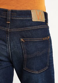 Jeans av denim med orange sömmar, en bältepatch av tan läder med en orange logotyp och en bakficka med ett sött mönster.