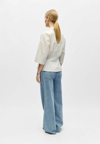 Femme aux cheveux blonds attachés en queue de cheval, portant une chemise blanche à manches mi-longues et un jean bleu à jambes larges, vue de dos sur un fond blanc uni.