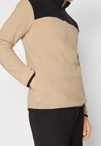 Sudadera de forro polar en beige con un panel superior negro, con un cuello alto y bolsillos laterales, textura suave y acentos de color en contraste.
