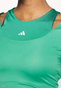 Primer plano de una persona que lleva dos camisetas deportivas verdes superpuestas, la exterior es más clara con detalles recortados en los hombros y un logo triangular blanco visible.