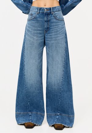 Weit geschnittene blaue Jeans mit hohem Bund, dezentem Ausbleichen und rohem Saum, getragen mit braunen Schuhen mit quadratischer Spitze.