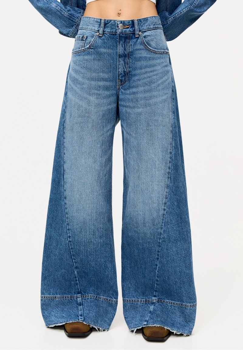 Jean en denim bleu taille haute à jambes larges avec un délavage subtil et un ourlet brut, porté avec des chaussures marron à bout carré.