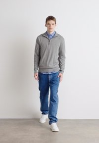 Sweat à zip gris sur une chemise bleu clair, associé à un jean bleu et des baskets blanches. Modèle debout devant un fond uni.