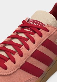 Κοντινή εικόνα αθλητικού παπουτσιού από σουέντ με ροζ επάνω μέρος, κόκκινες ρίγες και κορδόνια, μπεζ γλώσσα και σόλα από καουτσούκ, που δείχνει λεπτομερή ραφή και υφή.