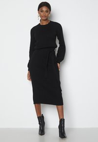 Set composto da un maglione nero a coste e una gonna midi, con vita a cravatta e maniche lunghe, abbinato a stivaletti neri lucidi.