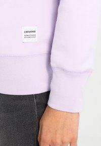 Lila sweatshirts har en mjuk textur, ribbade ärmmuddar och en märkesetikett som läser "CONVERSE REMASTERED ESSENTIALS" nära fållen.