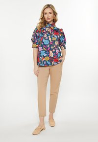 Bunte Blumenbluse mit kurzen Puffärmeln, hohem Kragen und Bindebändern, kombiniert mit beigen Slim-Fit-Hosen und hellen Loafers.
