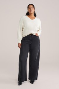 Pull blanc à col en V avec tissu texturé, associé à un jean noir taille haute et ample. Des bottines noires à talons complètent la tenue.