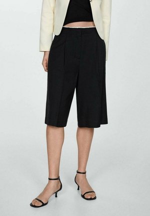 Persona que lleva pantalones cortos anchos negros hasta la rodilla, tacones negros con correa en el tobillo, top negro corto y blazer crema sobre un fondo liso.