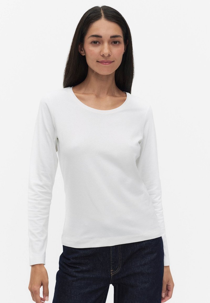 Woman with long dark hair wearing white long-sleeve crew neck shirt and dark blue jeans, standing against a plain white background.