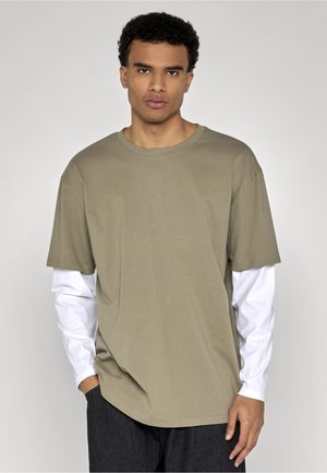 Tânăr care poartă un tricou larg verde măsliniu cu mâneci scurte peste o cămașă albă cu mâneci lungi și pantaloni închiși la culoare, stând în fața unui fundal gri.