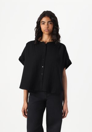 Camicia nera a maniche corte con bottoni in un tessuto strutturato, caratterizzata da un collo rotondo e una vestibilità ampia, abbinata a jeans scuri.