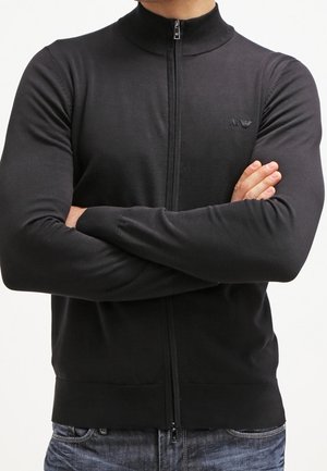 Mies pukeutuneena mustaan vetoketjulliseen pitkähihnaiseen neuleeseen, jossa on Armani-logo, kädet ristissä, yhdistettynä sinisiin farkkuihin.