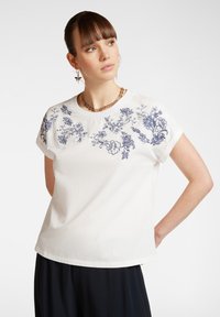 T-shirt blanc à manches courtes avec broderie florale bleue le long des épaules. Encolure ronde, coupe décontractée, matière en coton douce, texture minimaliste.