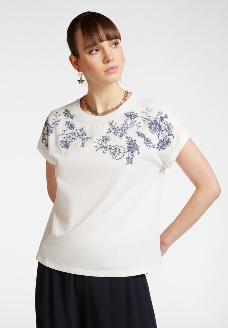 T-shirt blanc à manches courtes avec broderie florale bleue le long des épaules. Encolure ronde, coupe décontractée, matière en coton douce, texture minimaliste.