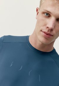 Krátký rukáv, přiléhavé sportovní triko v tmavě modré látce. Má hladkou texturu a jemný detail loga na přední straně. Kulatý výstřih.