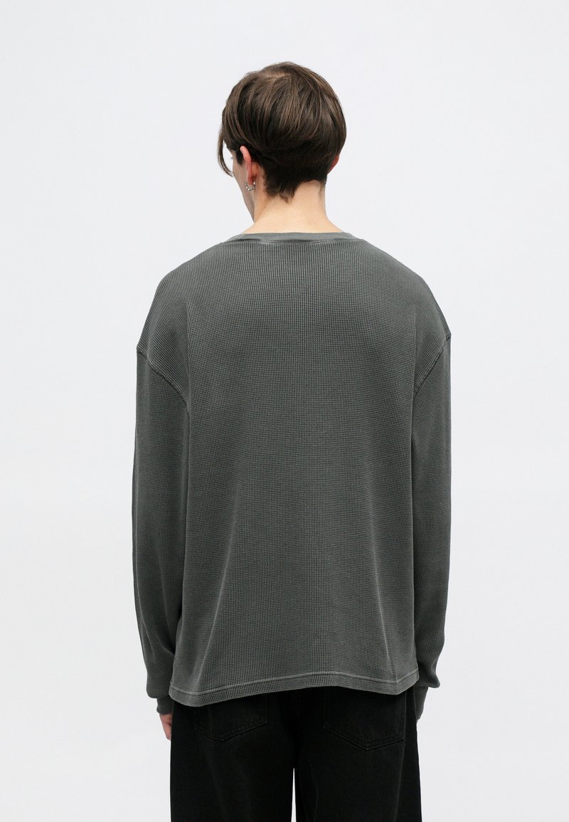 Übergroßer dunkelgrauer Strickpullover mit gerippter Textur, fallenden Schultern und weitem Ausschnitt. Die langen Ärmel reichen bis zu den Handgelenken.