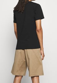 Black cotton t-shirt with short sleeves, viewed from the back, paired with tan knee-length shorts made of a lightweight fabric.