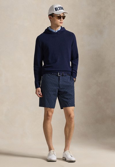 Muž v tmavě modré mikině s kapucí, vzorovaných tmavě modrých šortkách, bílém opasku, bílé čepici s tmavým nápisem, slunečních brýlích a bílých botách stojící na neutrálním pozadí.