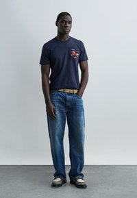 Hombre de pie con las manos en los bolsillos, vestido con una camiseta azul marino con un gráfico rojo en el bolsillo, vaqueros azules, cinturón color canela y zapatos bicolor.