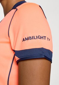 Korte mouwen sportshirt in helder koraal met donkere blauwe accenten. Voorzien van de opdruk "AMBI LIGHT tv" op de mouw. Gelaagd materiaal.