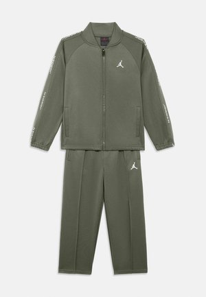 Survêtement vert olive en tissu doux, comprenant une veste zippée avec poches latérales et un pantalon assorti. Détails de logo blanc sur les deux pièces.