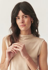 Žena s dlouhými tmavými vlasy, která má na sobě bezrukávový béžový top, zlaté náušnice a náramek, drží ruce kolem obličeje na neutrálním pozadí.